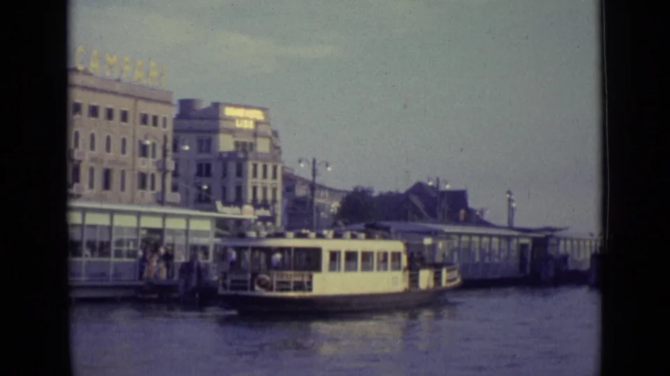 A 1962 Venice canal-side dock scene with a boat at the pier, people near the landing, and a Campari sign above the waterfront buildings.
