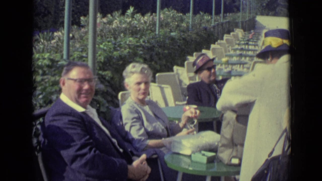 Adults seated at green café tables on a sunny terrace with rows of chairs behind them.