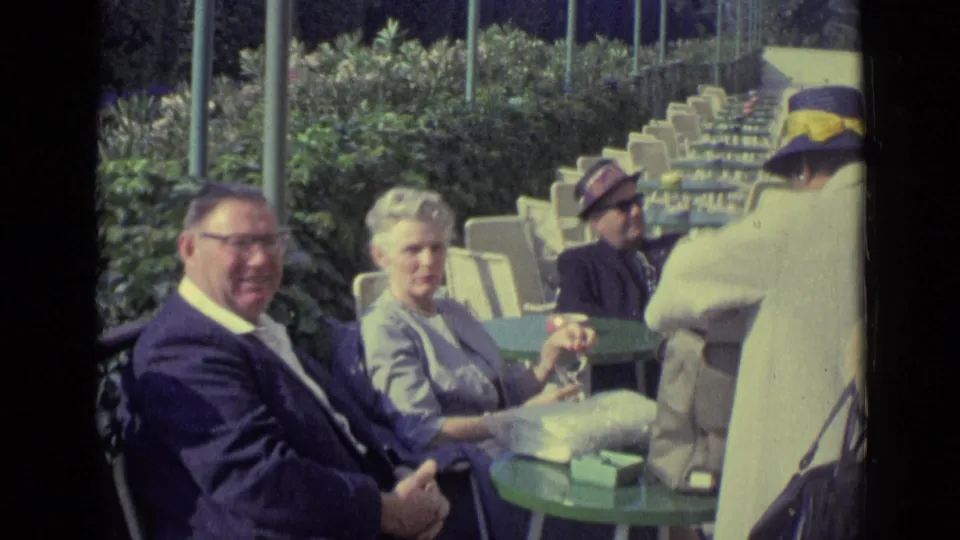 1962 Venice home-movie frame showing adults at outdoor café tables, including a man in glasses looking toward the camera.