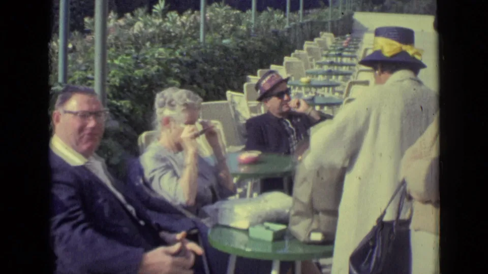1962 Venice café scene with adults at green tables, including a woman drinking and a man in glasses facing the camera.