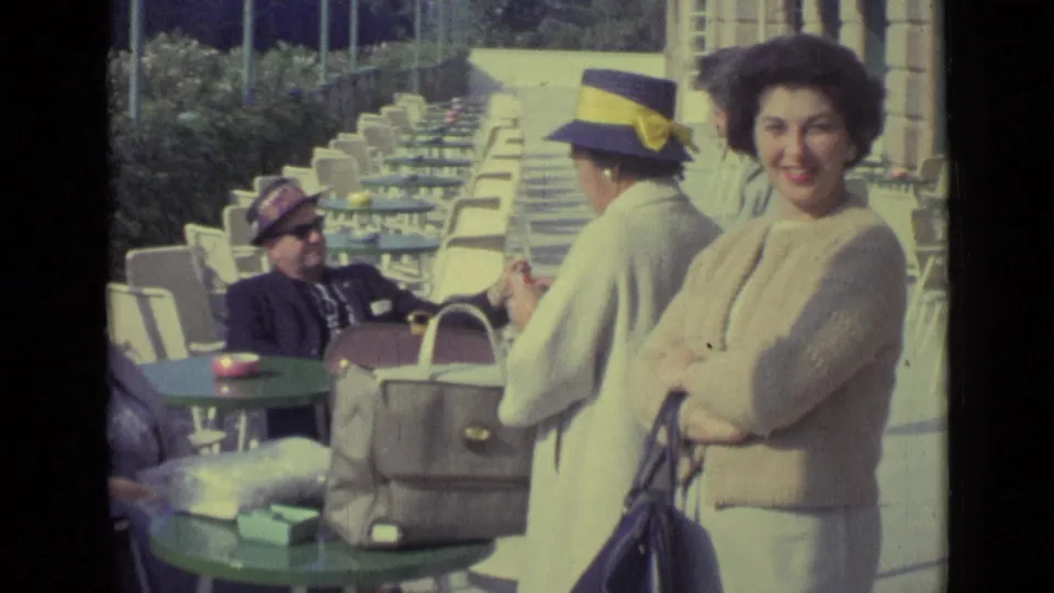1962 Venice home-movie frame with a smiling woman beside outdoor café tables, bags, and seated adults.
