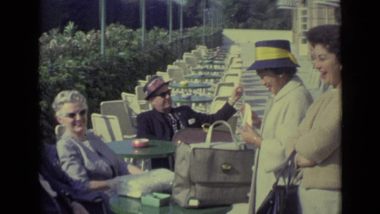 Adults seated at green cafe tables near striped beach cabanas in bright sun.