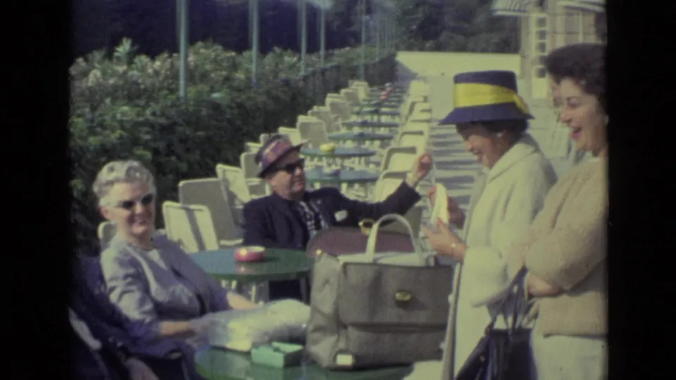 1962 Venice resort home-movie frame showing adults at green outdoor cafe tables beside striped beach cabanas.