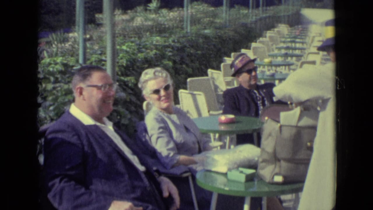Man and woman seated at a green cafe table with cabanas and chairs behind them.