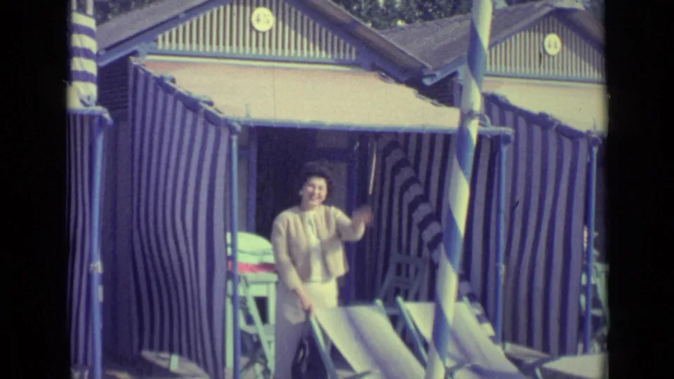 1962 Venice home-movie frame of a woman in light clothing standing before a striped beach cabana.