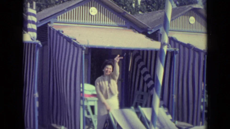 1962 Venice resort frame showing a woman smiling and waving at the camera from a striped beach cabana doorway.