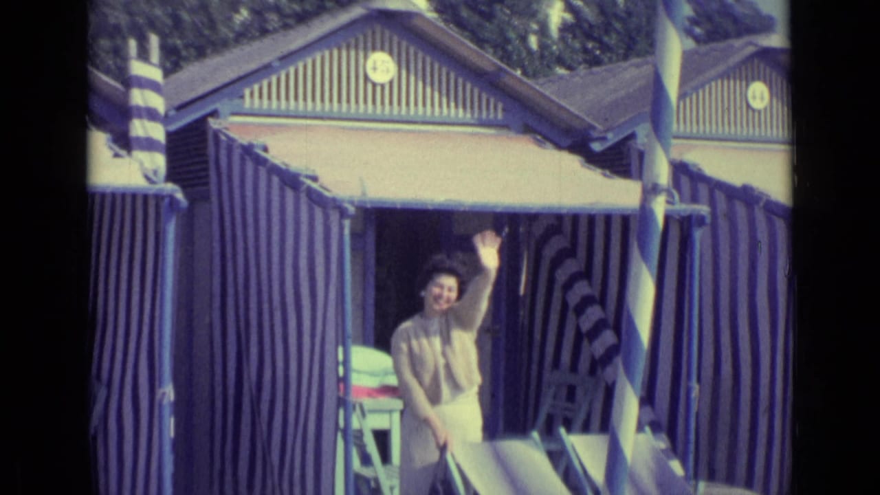 In 1962 Venice, Italy footage, a woman waves from a blue and white striped beach hut beside deck chairs.