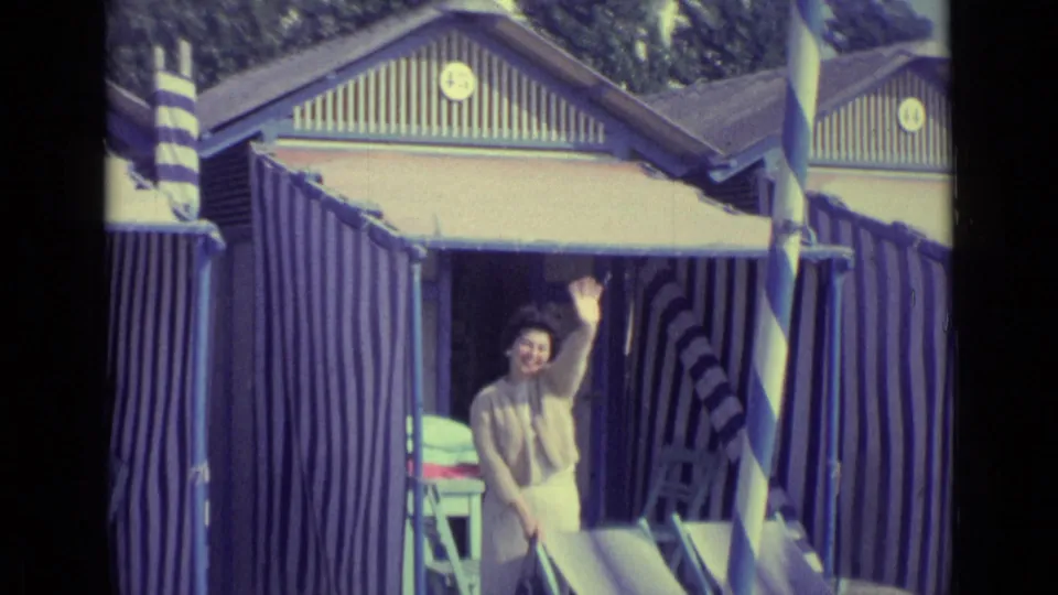In 1962 Venice, Italy footage, a woman waves from a blue and white striped beach hut beside deck chairs.