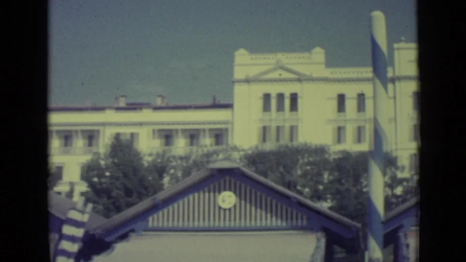 1962 Venice resort scene with a striped pole, beach hut roof, trees, and a large pale building in the background.
