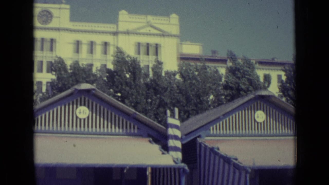 1962 Venice frame with striped beach huts in the foreground and a pale classical building seen above trees.