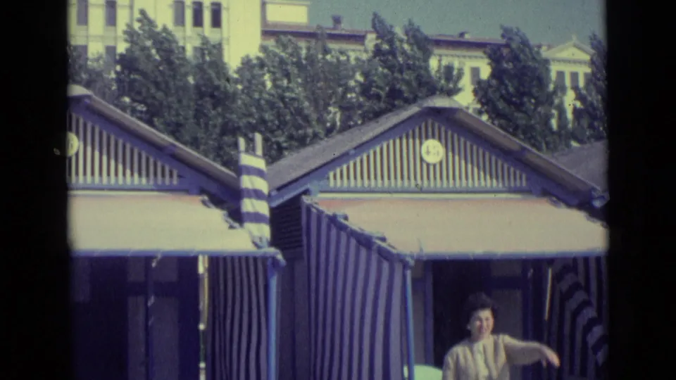 1962 Venice home-movie frame showing striped beach huts and awnings as a woman appears near the lower edge.