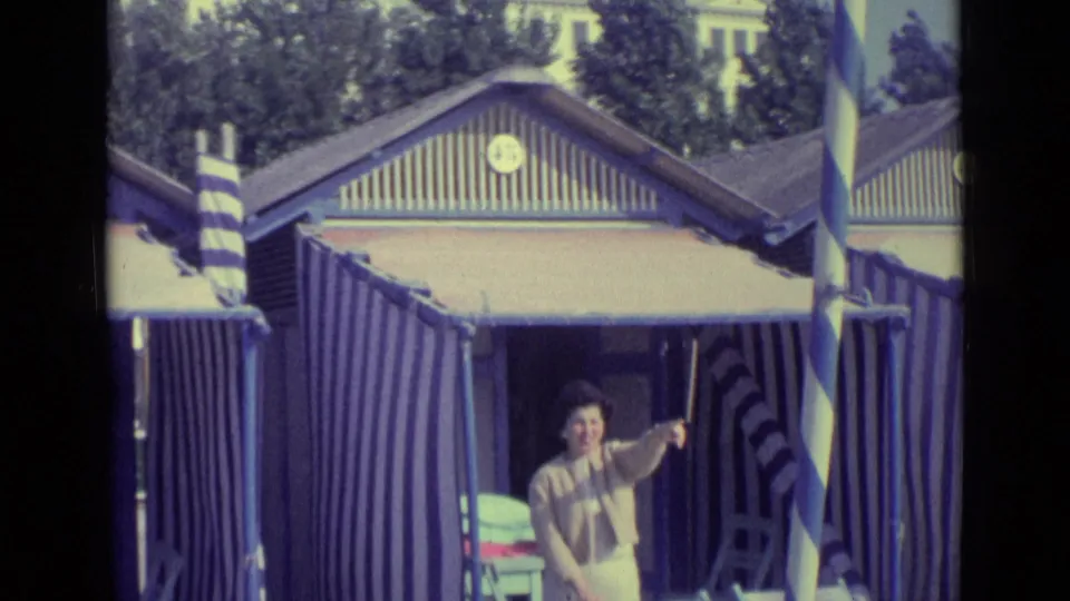 1962 Venice frame showing a woman smiling and pointing near striped beach huts on a sunny seaside promenade.