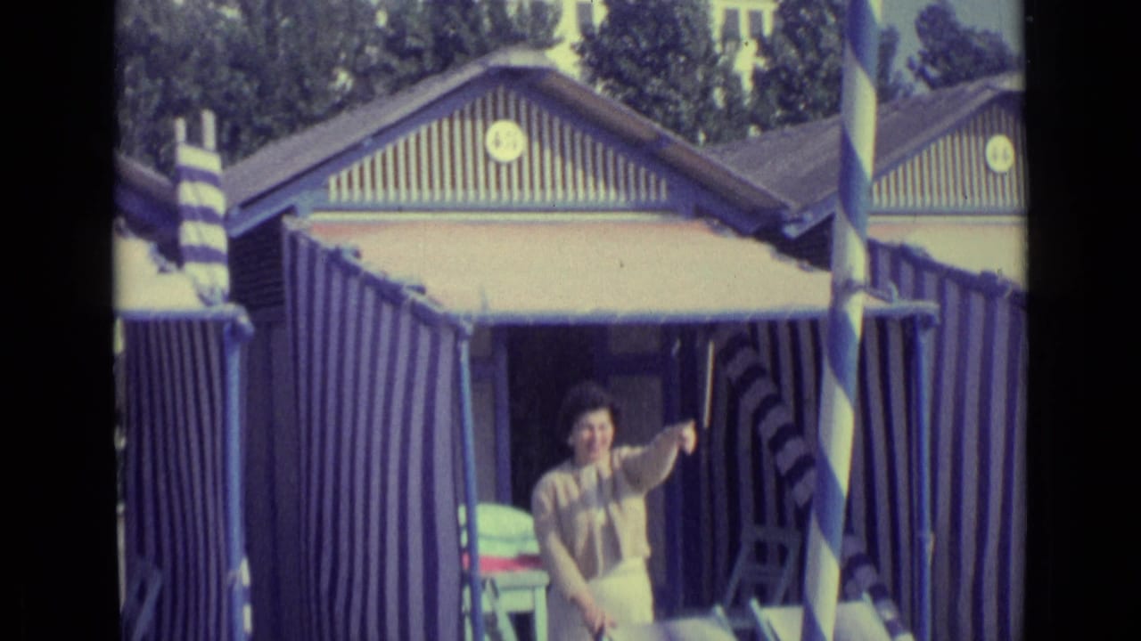 1962 Venice beach view with a woman gesturing from a blue and white striped hut doorway, chairs visible at the entrance.