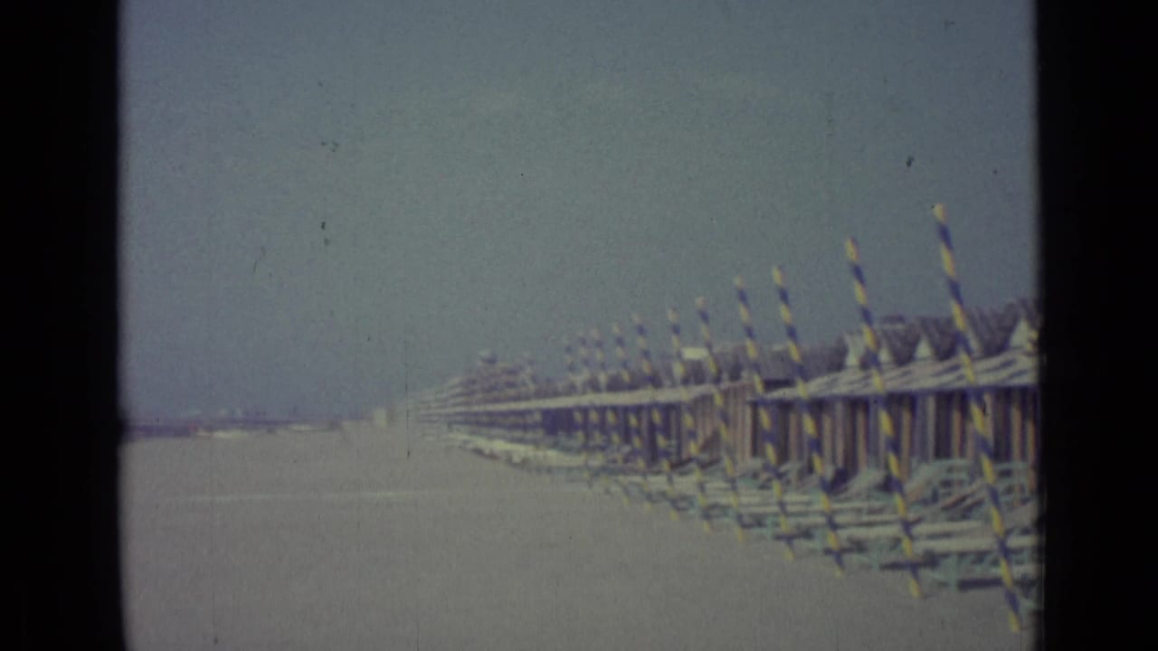 A long line of striped beach huts and poles stretches along the sandy Venice shoreline under a hazy sky.