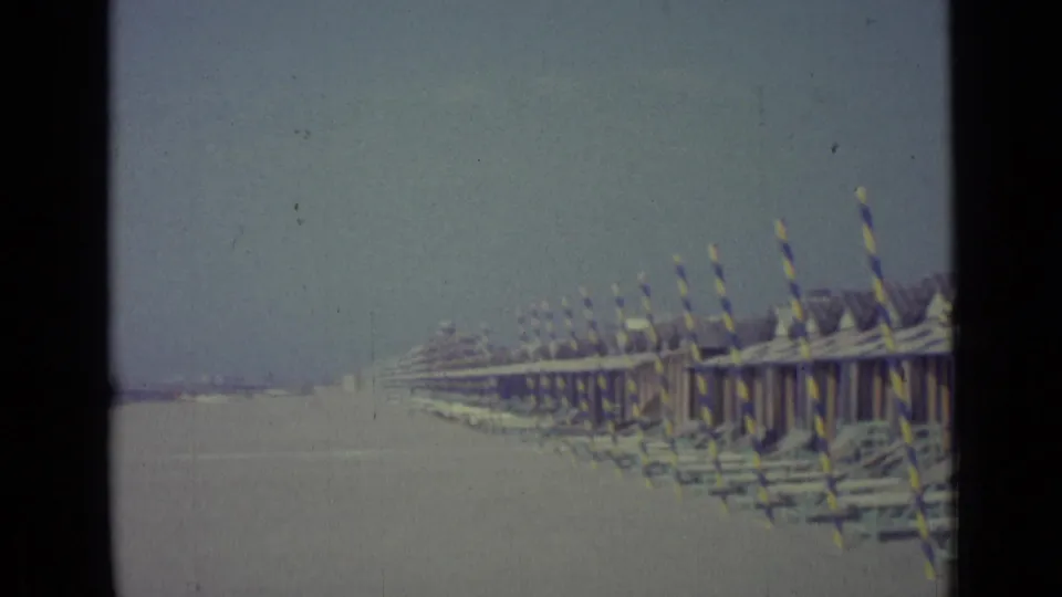 A long line of striped beach huts and poles stretches along the sandy Venice shoreline under a hazy sky.