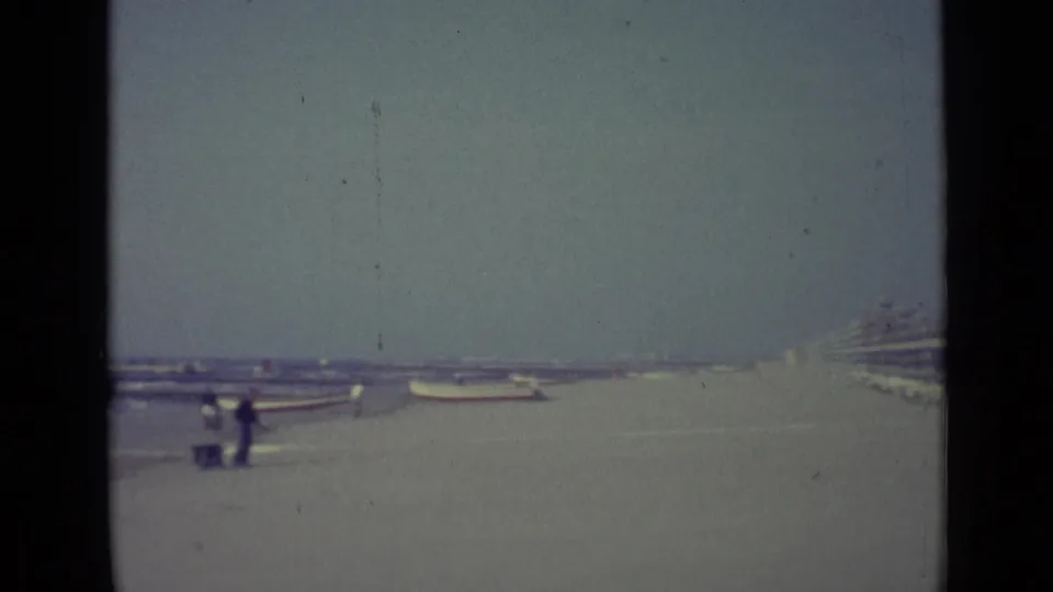 Wide 1962 Venice beach view with small boats on the sand and distant figures walking near the waterline.
