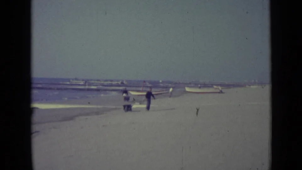 Two distant figures walk across a sandy Venice beach, with small boats near the shoreline and a hazy sea horizon beyond.