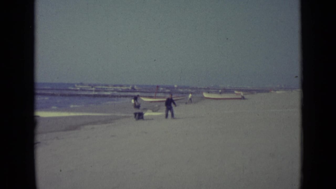 Man in dark clothing walks on a sandy beach near boats and a person with a wheelbarrow.