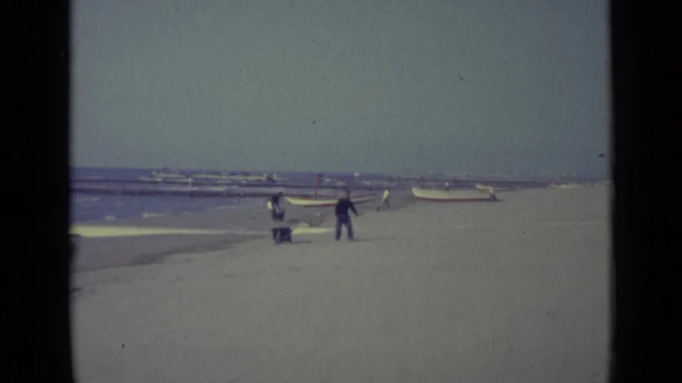 1962 Venice beach view with a man carrying a pole, small boats near the shoreline, and another person beside a wheelbarrow.
