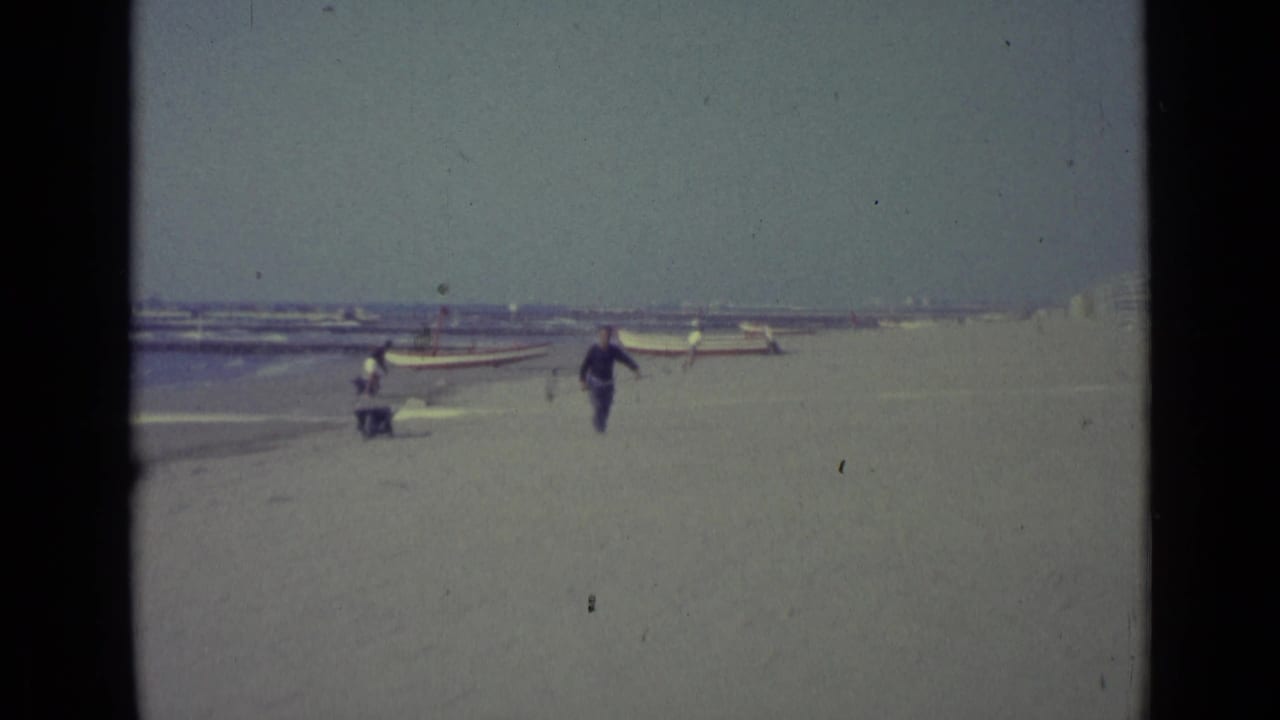 On a Venice beach in 1962, a man in dark blue clothing carries a pole while small boats and distant people line the shore.