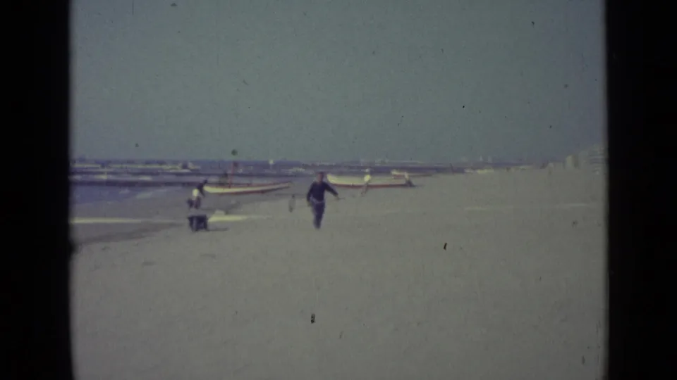 On a Venice beach in 1962, a man in dark blue clothing carries a pole while small boats and distant people line the shore.