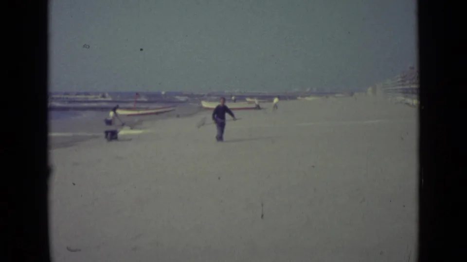 1962 Venice shoreline scene showing a man with a pole in the sand, a wheelbarrow near the surf, and small boats pulled up behind him.