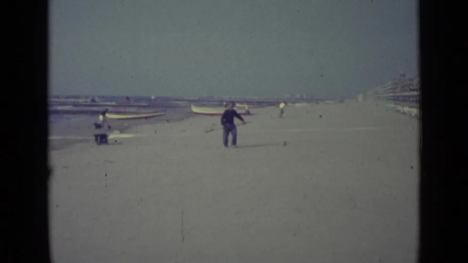 The 1962 Venice beach view frames the man with a pole nearer to camera, with boats, distant figures, and waterfront structures in the background.