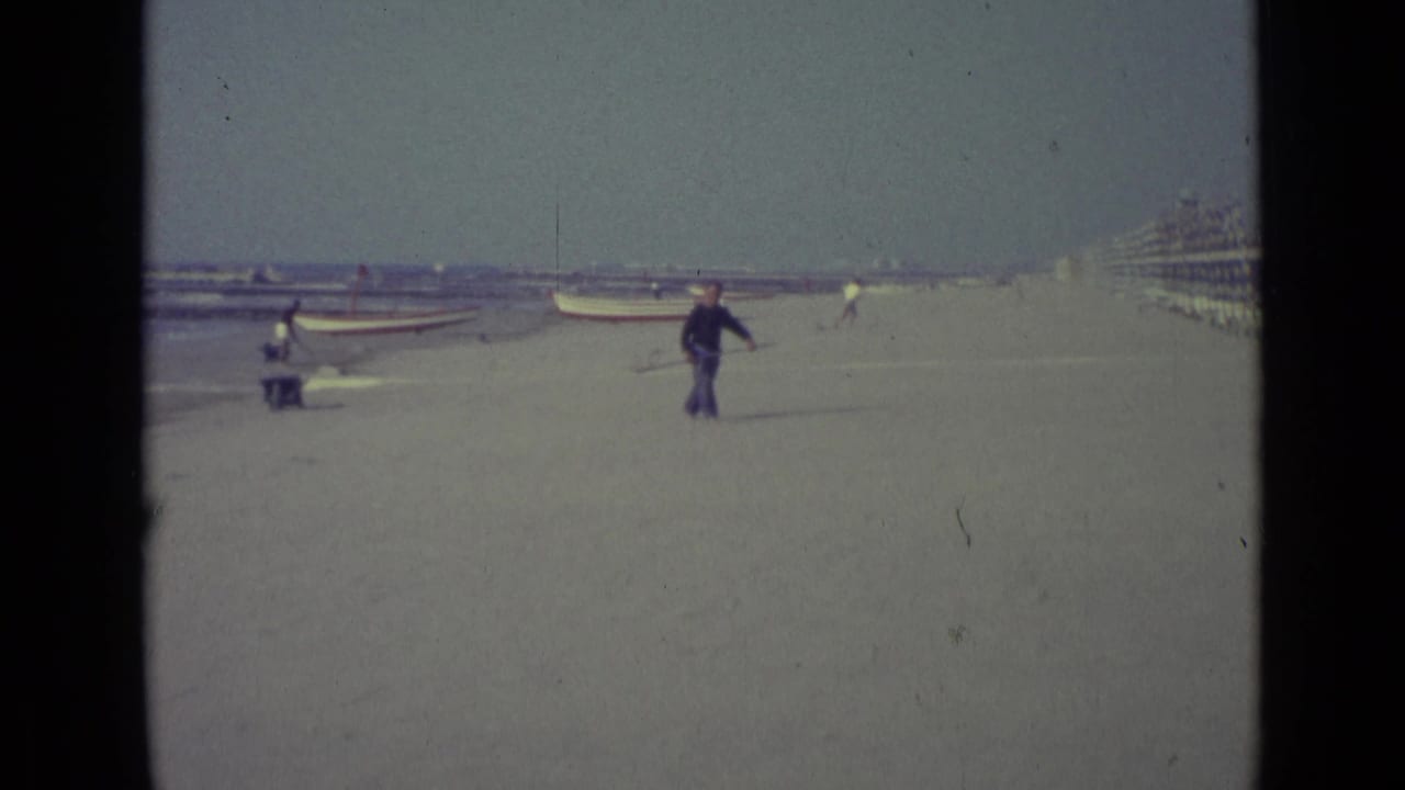 Man walks across a wide beach with a net near boats and striped cabanas.