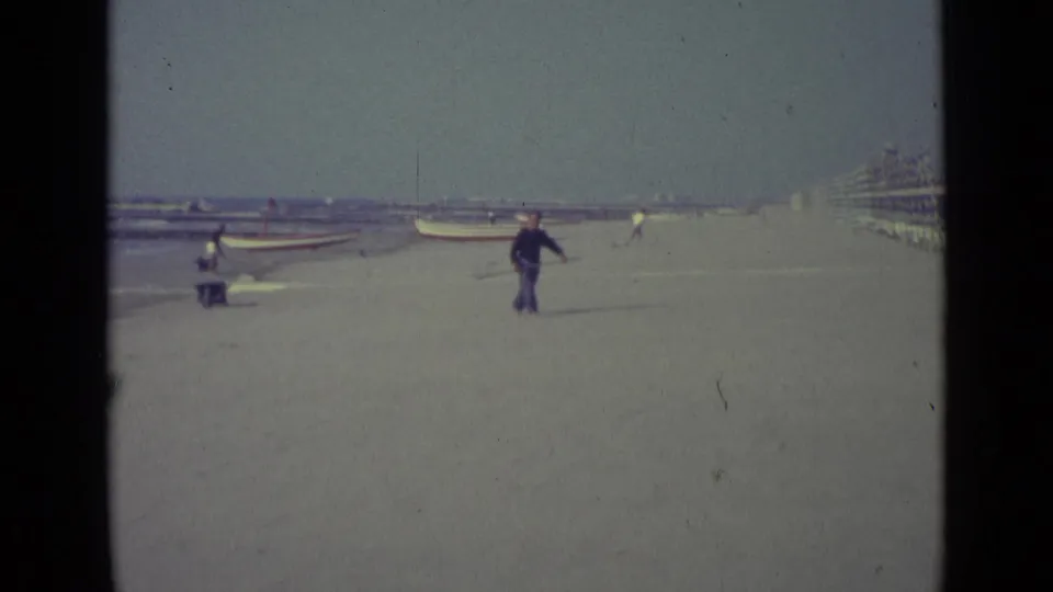 1962 home-movie frame from Venice, Italy showing a man crossing a sandy beach with a long-handled net near boats and cabanas.