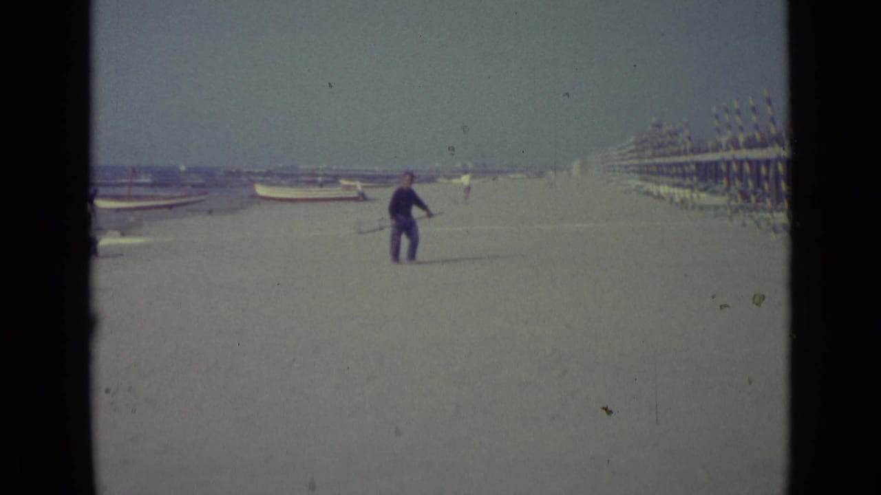 Man with a long net walks past cabanas on a broad sandy beach.
