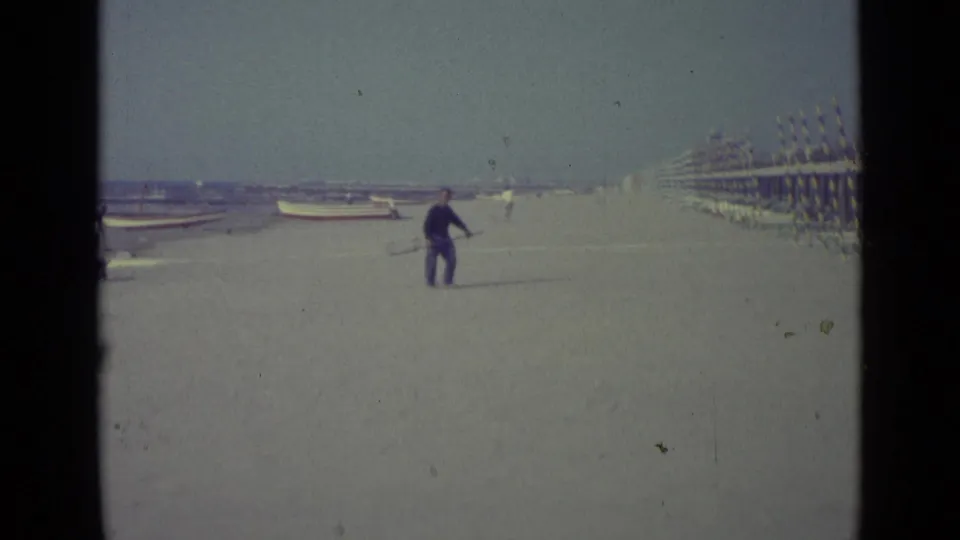 The man continues across the Venice beach with a long-handled net, with small boats, cabanas, and distant figures visible.
