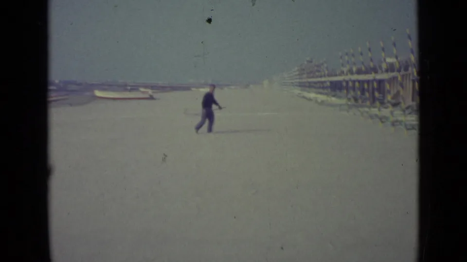 A distant man with a long-handled net walks near rows of striped cabanas on the 1962 Venice beach scene.