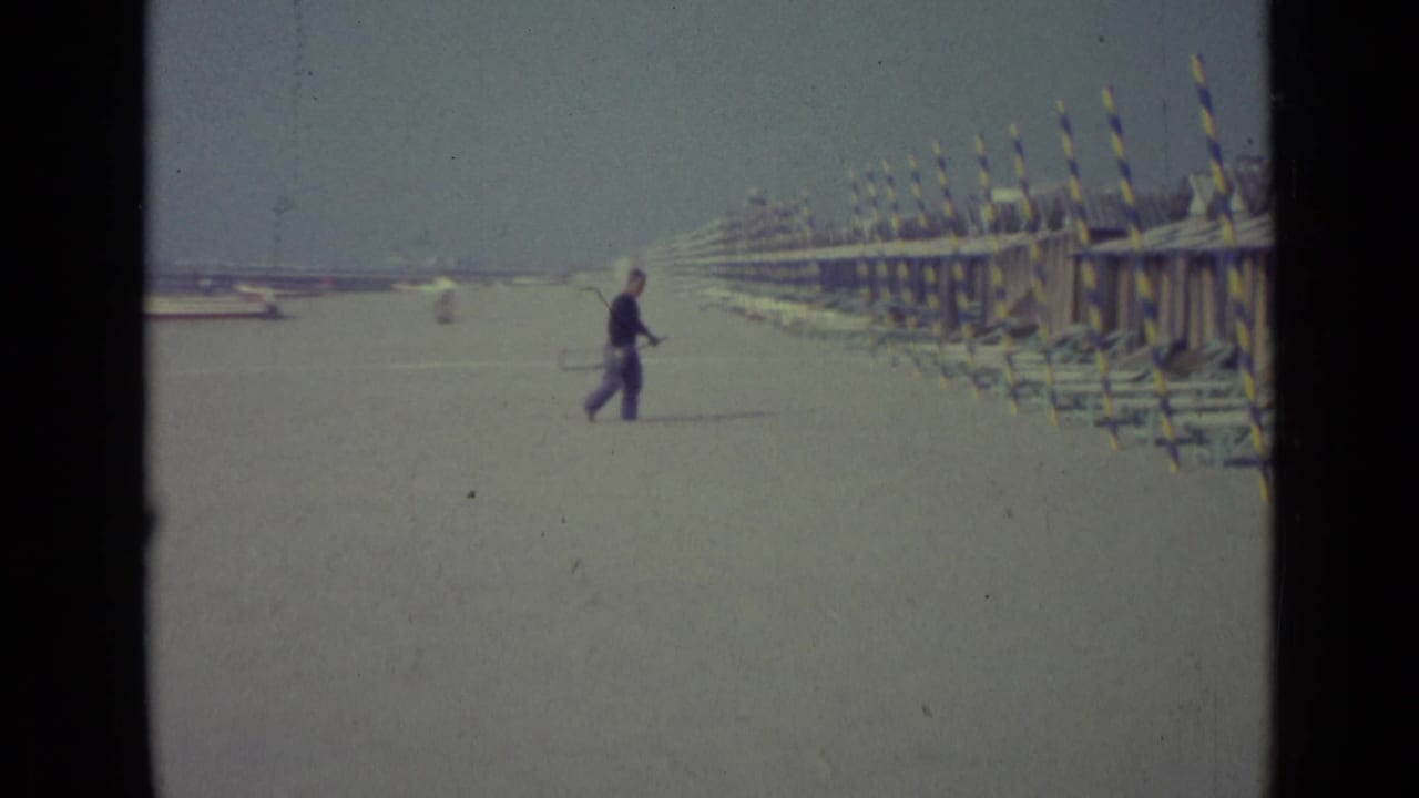 1962 Venice beach view with a man crossing open sand near long rows of striped huts or cabanas and lounge chairs.