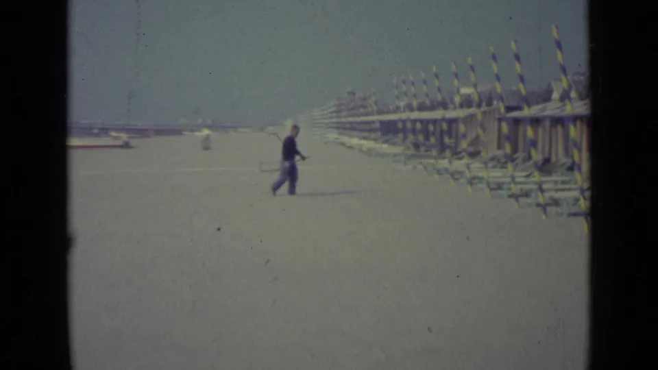 1962 Venice beach view with a man crossing open sand near long rows of striped huts or cabanas and lounge chairs.