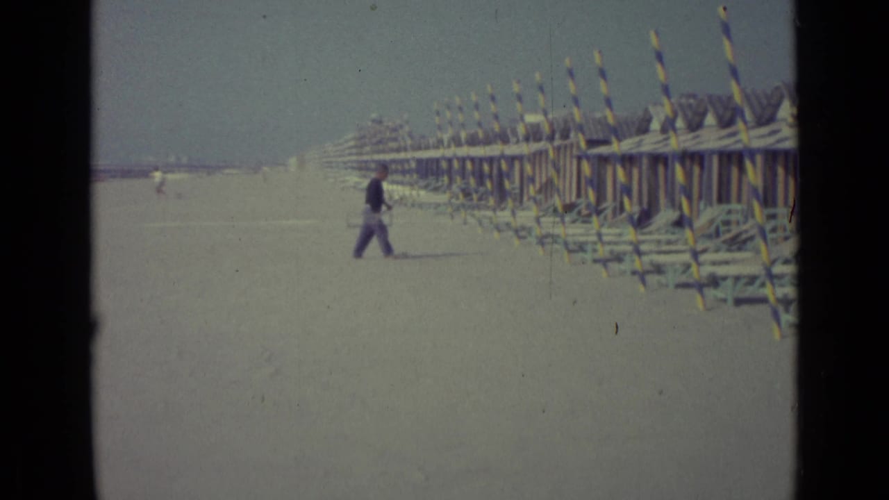 Distant man on a Venice beach in 1962, moving near rows of striped cabanas and stacked beach furniture.
