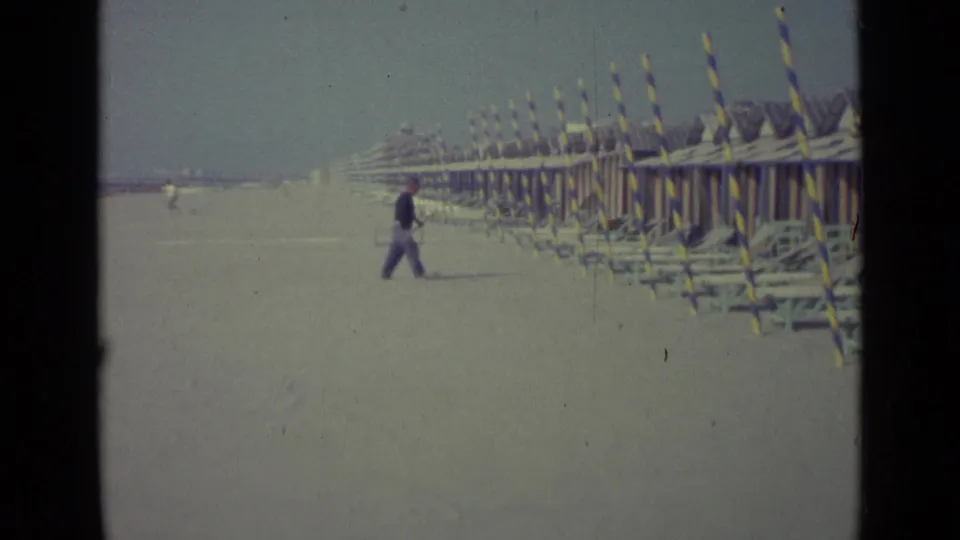 Distant man on a Venice beach in 1962, moving near rows of striped cabanas and stacked beach furniture.