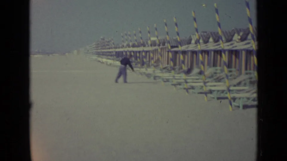 1962 Venice beach scene with a man handling a long pole near aligned chairs and striped seaside cabanas.