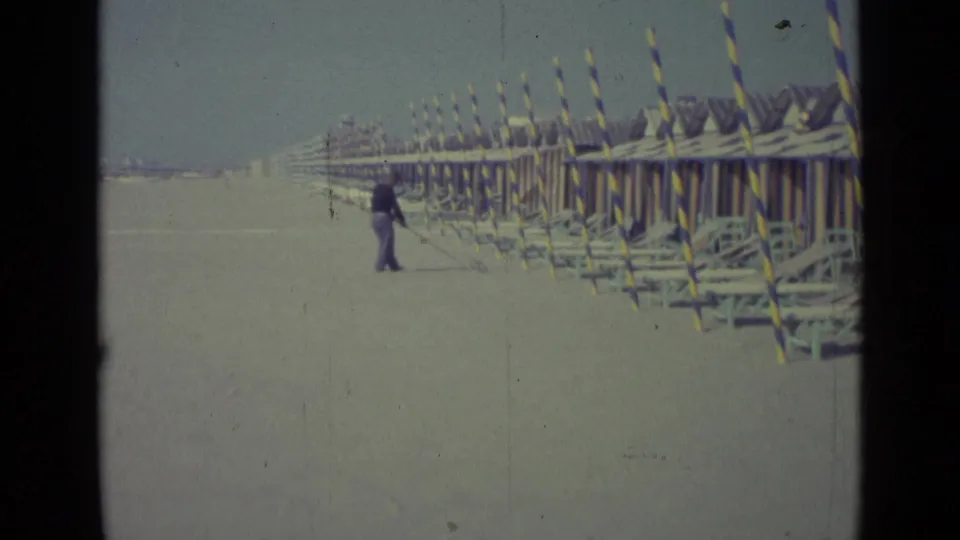 Man on a 1962 Venice beach holding a long pole near orderly rows of cabanas and lounge chairs.