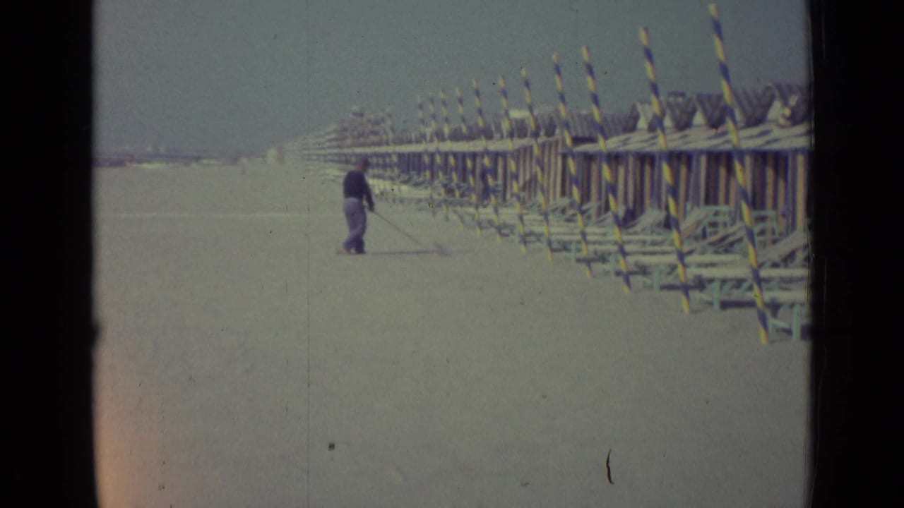 Man sweeping pale sand beside rows of striped beach structures.