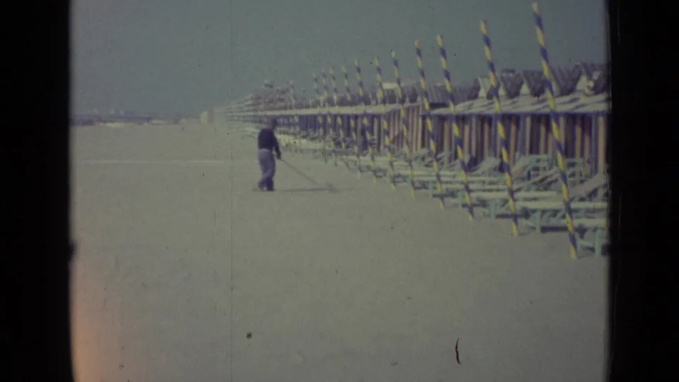 1962 Venice, Italy 8mm frame showing a distant man sweeping a quiet beach near striped beach huts or poles.
