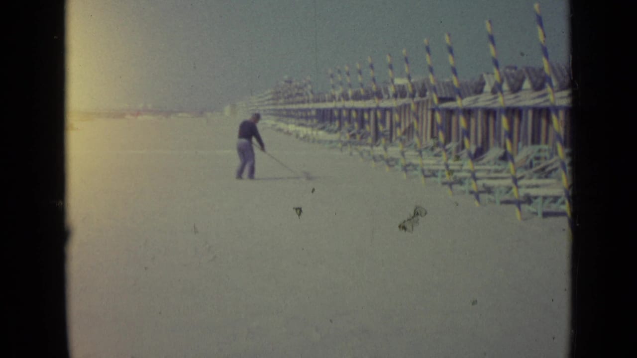 Distant man cleaning sand near striped beach huts under a hazy sky.