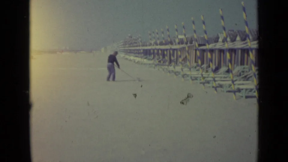 Distant beach worker or visitor sweeps sand on a Venice beach in 1962, with striped structures receding along the shore.