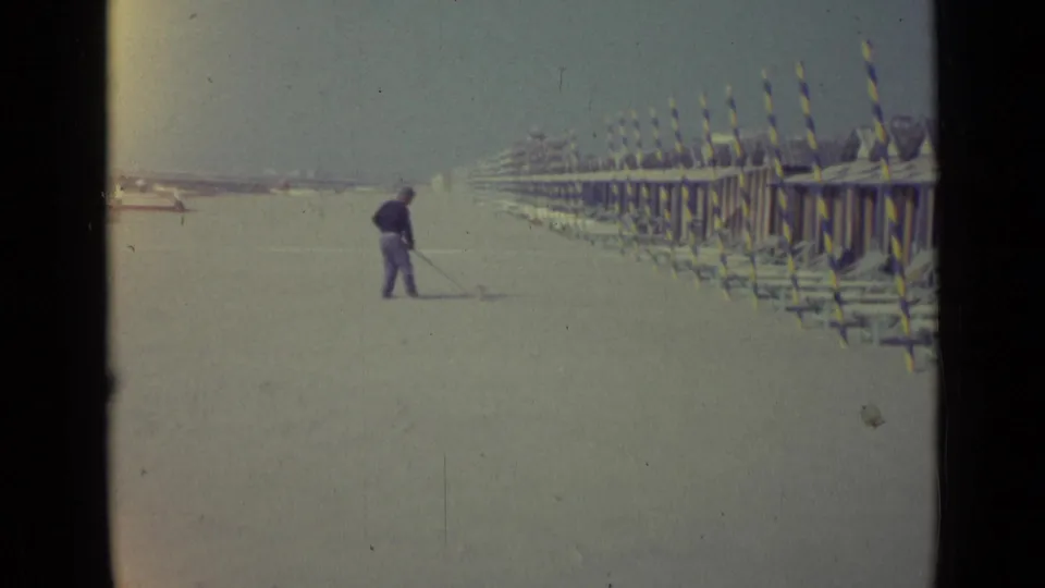 1962 home-movie frame of a man sweeping a quiet Venice beach, with striped huts or poles lining the right side.