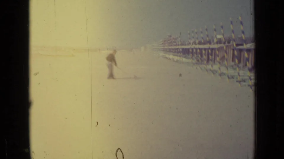 Faded 8mm frame from Venice, Italy in 1962 showing a man sweeping sand beside rows of striped beach structures.