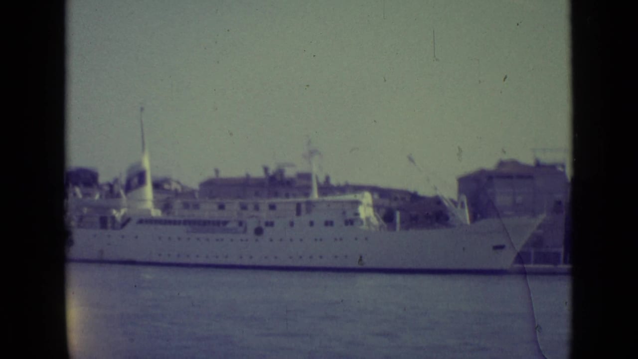 Docked white passenger ship beside waterfront buildings, partly framed by dark film borders.