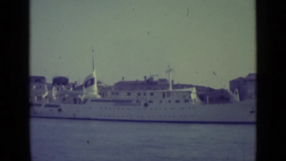 Faded 8mm frame of a white passenger ship in Venice, showing its decks, mast, and waterfront buildings beyond.