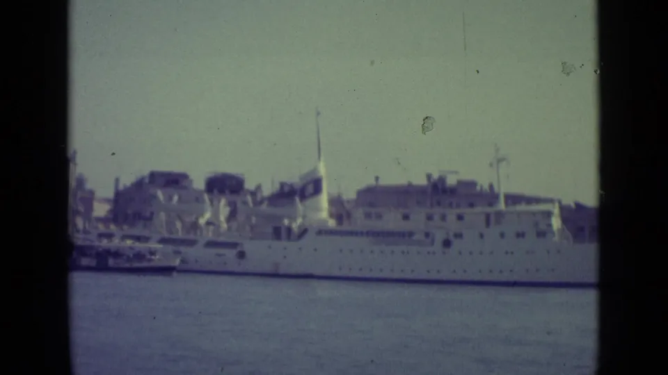 1962 8mm view of a large white passenger ship docked across canal water in Venice, with historic waterfront buildings in the background.