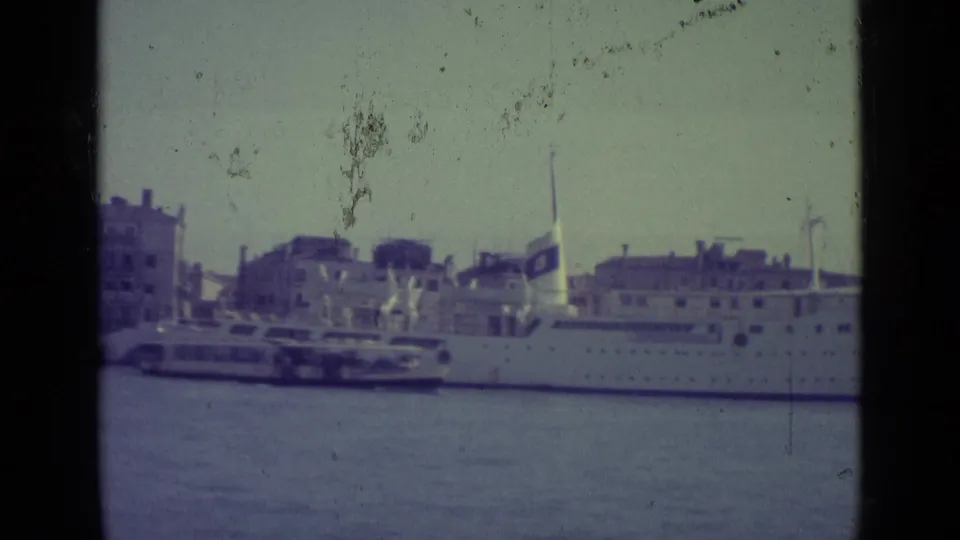 Venice canal view from 1962 showing a docked white ship, smaller ferry traffic, and waterfront buildings in soft 8mm color.