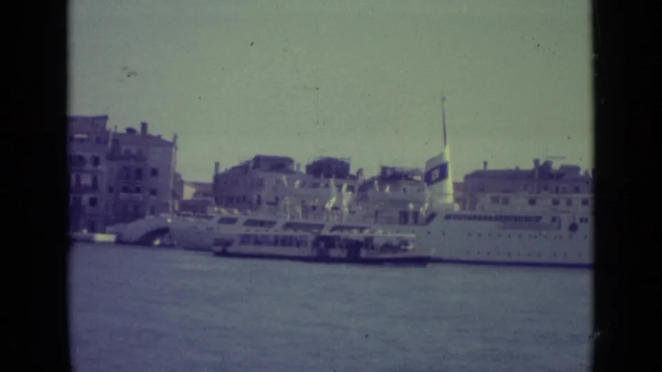 1962 Venice home-movie frame with canal water, waterfront buildings, a small ferry, and part of a large docked passenger ship.