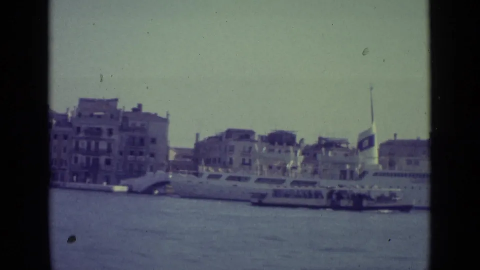 Final 1962 8mm frame looking across Venice canal water toward waterfront buildings, small boats, and the edge of a large docked ship.